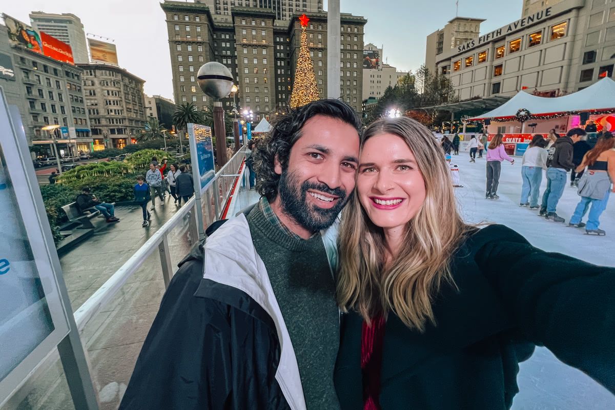 A dark-haired man and a light-haired woman put their heads together and smile as they take a selfie on the Union Square Ice Rink, with a giant Christmas tree behind them.
