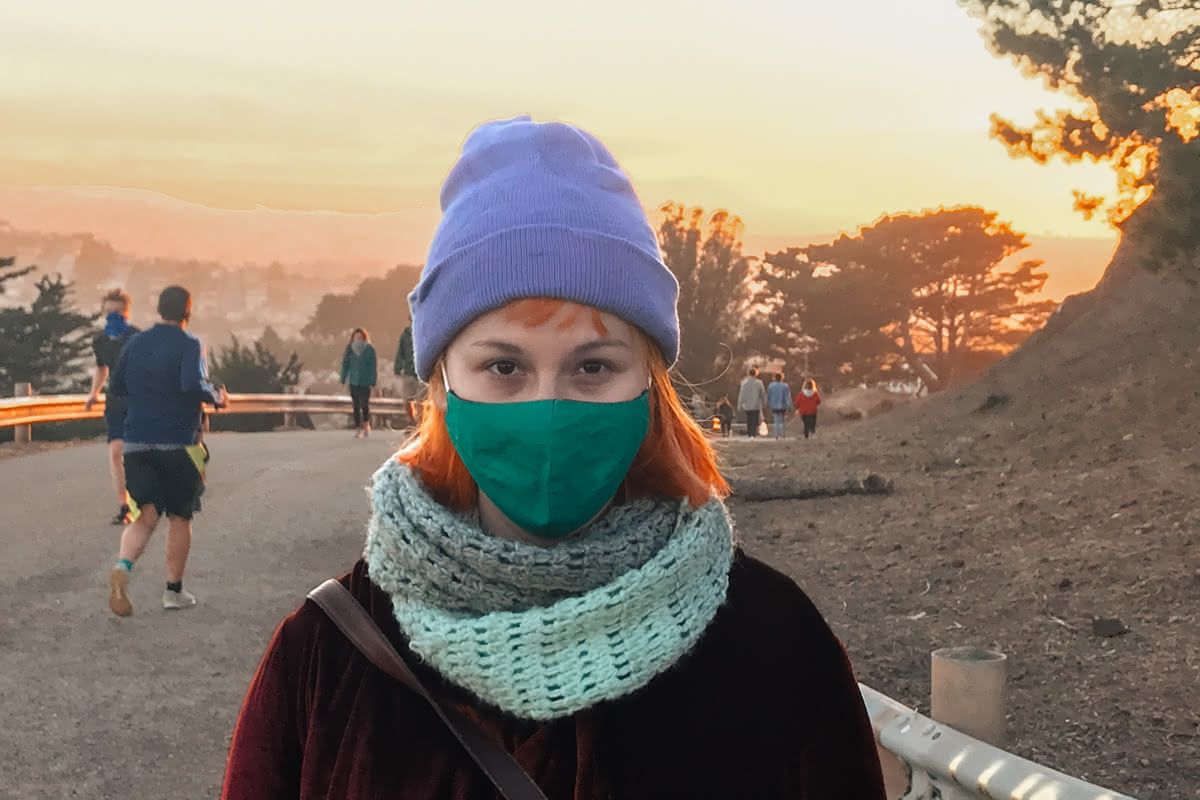 A young woman with orange hair wearing a purple beanie, turquoise face mask, and teal knit scarf stands facing the camera on a hilltop with an orange sunset in the background.