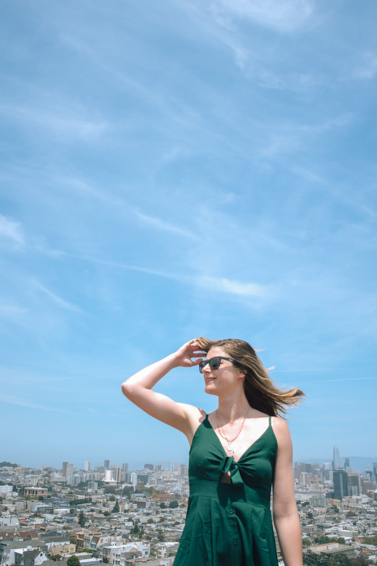 A young woman in a green dress stands on a hilltop in front of a San Francisco cityscape on a sunny day, hair blowing in the wind.