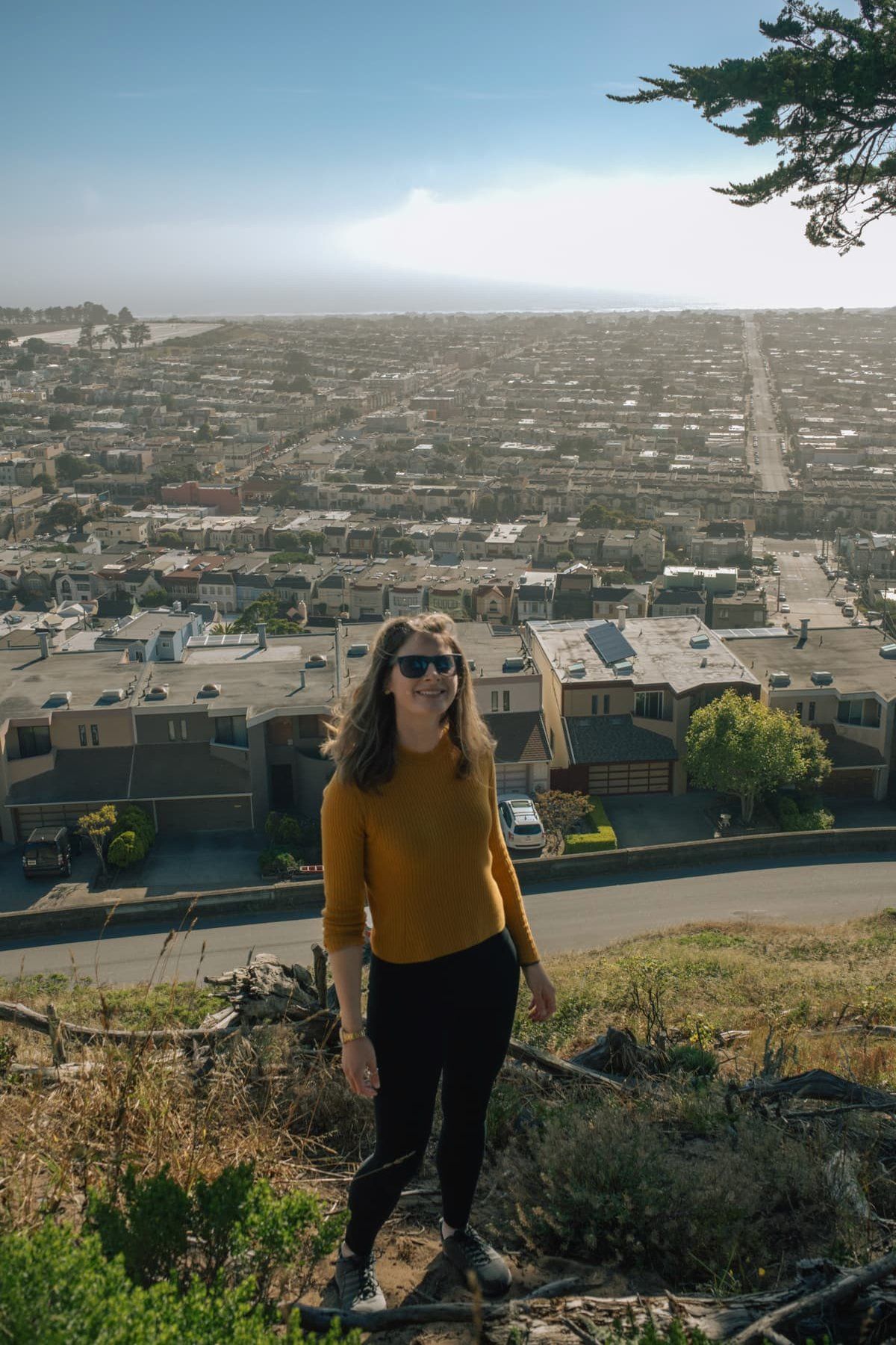 A young light-haired woman in black pants and a yellow shirt smiles at the camera as she stands on a hillside with a view of San Francisco behind her.