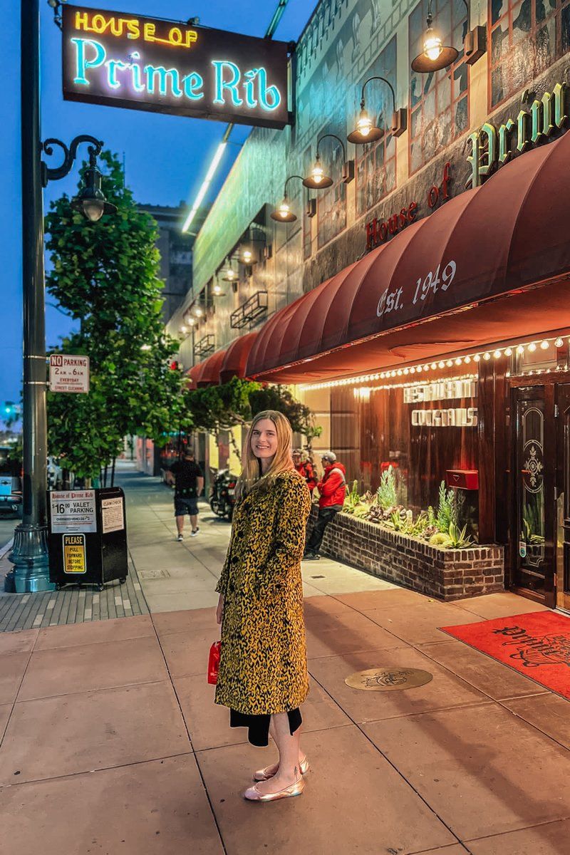 A woman in a leopard-print coat smiles over her shoulder while standing in front of the House of Prime Rib at dusk.