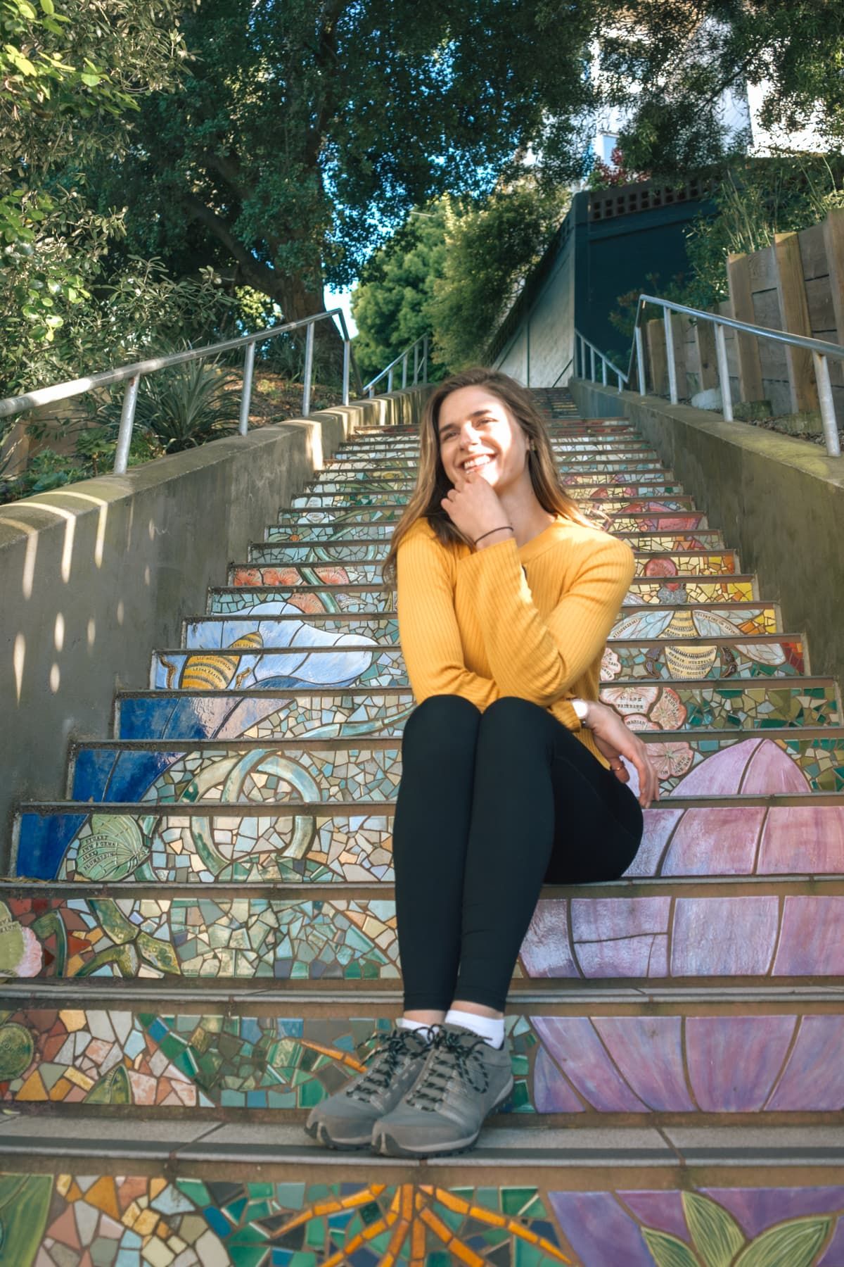 A young light-haired woman in black pants and a yellow shirt smiles at the camera as she sits on a set of stairs decorated with mosaics.