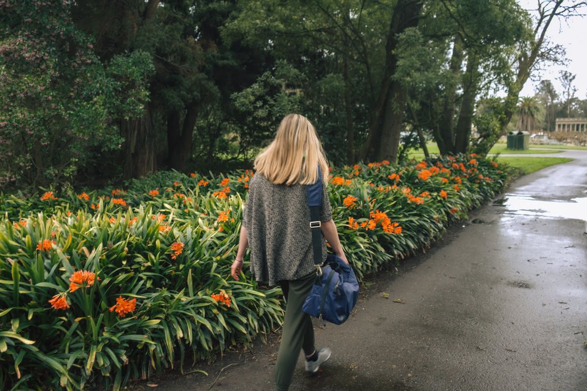 A young light-haired woman see from behind walking away with a blue duffel bag on her shoulder in a San Francisco park.